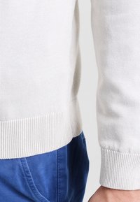 Gros plan sur une personne portant un pull en tricot blanc et un pantalon bleu, mettant en évidence l'ourlet et les poignets côtelés du pull.