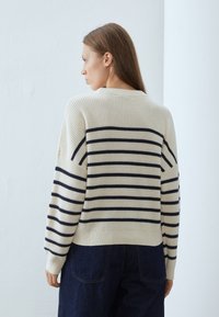 Kremfarget strikket genser med marineblå horisontale striper, ribbede kantlinjer i hals og armer, og en avslappet passform. Myk tekstur, detaljerte sømmer.