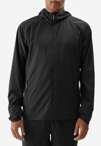 Czarna, lekka kurtka z kapturem, zapinana na zamek z przodu i z kieszeniami po bokach. Gładka faktura, minimalna ilość szwów oraz kontrastujące zielone akcenty zamka.