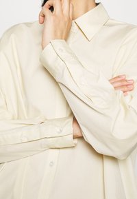 Chemise de couleur crème avec un col classique, des manches longues et des poignets à boutons ; tissu lisse avec une texture minimale et aucun motif visible.