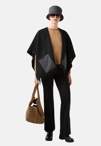 Poncho noir avec des bords frangés et deux poches noires brillantes, associé à un pull beige, un pantalon noir, un sac à épaule marron et un chapeau seau gris.