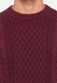 Suéter tricotado em burgundy com um padrão de cabos em diamante texturizado e gola redonda canelada. O tecido aparenta ser suave e volumoso.