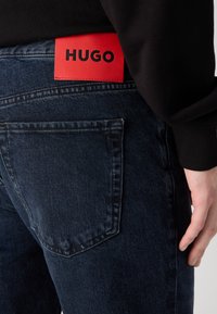 Tummansiniset farkut, joissa on erottuva punainen nahkatalvi "HUGO" oikean takapussin yläpuolella, sisältäen tavanomaisen ompelun ja taskusuunnittelun.