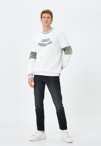 Sudadera blanca con el texto "LEADER GENERATION", rayas verdes y grises en las mangas, combinada con jeans oscuros y zapatillas blancas.
