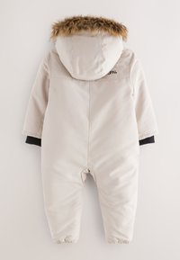 Beige insulated snowsuit with a fur-lined hood, elastic cuffs, and dark knit accents. Features minimal branding on the back.