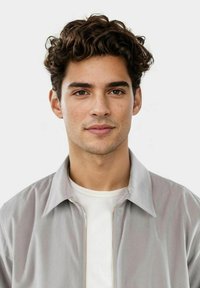 Grijze cordjack met een lichte textuur, geopend gedragen over een wit shirt. Het onderwerp heeft kort, krullend haar en een neutrale gezichtsuitdrukking.