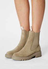 Beige Wildleder-Stiefeletten mit elastischen seitlichen Einsätzen, dicken Gummisohlen und genähten Details. Schlaufen-Überzieher an der Rückseite.
