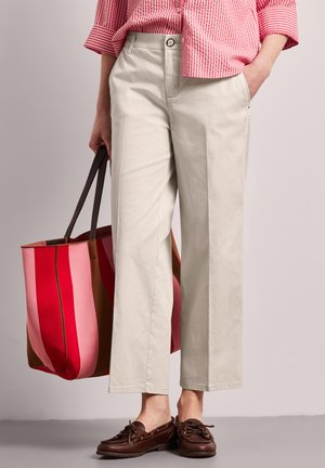 Femme portant un pantalon large beige, une chemise rayée rouge, et des mocassins marron, tenant un grand sac cabas rayé rouge et rose.