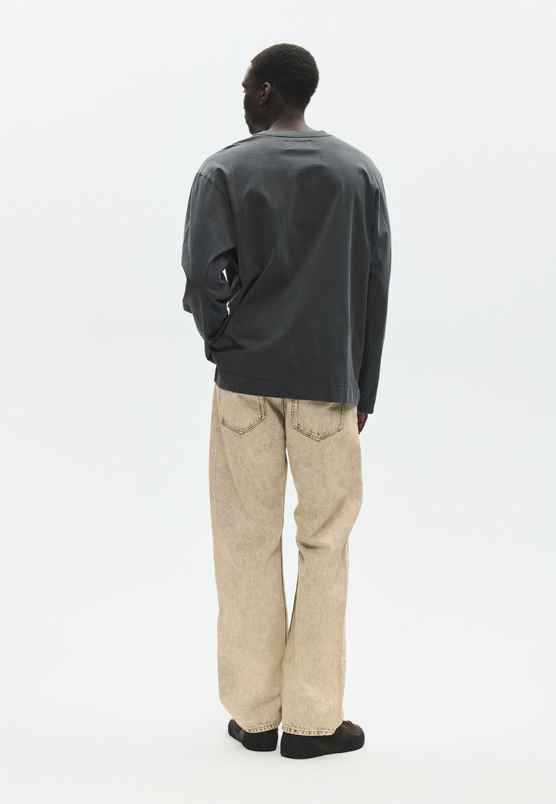 Ein Mann, von hinten zu sehen, trägt ein dunkelgraues Langarmshirt, beige weite Jeans und schwarze Schuhe vor einem einfarbigen hellen Hintergrund.