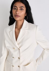 Off-white blazer with a double-breasted design, gold hardware accents, notched lapels, and two front pockets. Smooth fabric texture.