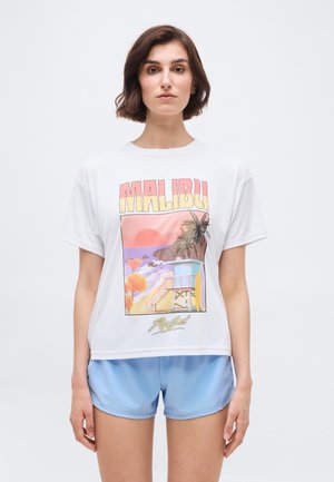 Γυναίκα που φοράει λευκό T-shirt με στάμπα "Malibu" με παραθαλάσσια σκηνή και ανοιχτό μπλε αθλητικό σορτς, στέκεται μπροστά σε απλό φόντο.