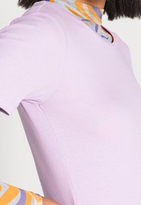 Fialový žebrovaný top s krátkým rukávem a kulatým výstřihem; vrstvený pod barevným vzorovaným topem s vysokým límečkem s oranžovými a modrými víry.