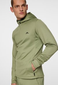 Huppari, jossa on huppu ja vaaleanoliivinvihreä väri, valmistettu sileästä kankaasta. Ominaisuuksiin kuuluvat puoli-vetoketju, sivutasku ja musta logo-ostos.