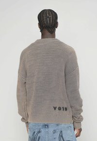 Übergroßer, gerippter Strickpullover in Taupe mit Rundhalsausschnitt. Verfügt über überschnittene Schultern und den Schriftzug "V015" in der Nähe des Saums.