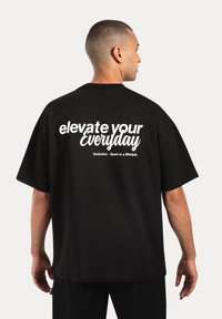 Sort oversized t-shirt lavet af bomuld, med hvid tekst "elevate your Everyday" på ryggen, med en afslappet pasform og korte ærmer.