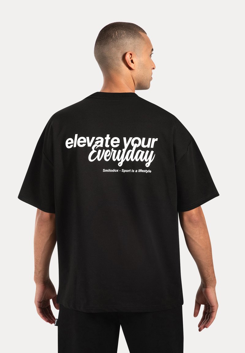 Sort oversized t-shirt lavet af bomuld, med hvid tekst "elevate your Everyday" på ryggen, med en afslappet pasform og korte ærmer.