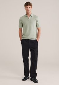 Jonge man staand met handen in zijn zakken, gekleed in een lichtgroene polo met korte mouwen, zwarte broek en zwarte nette schoenen, tegen een beige achtergrond.