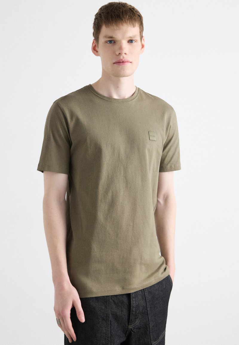 Tricou din bumbac de culoare verde măslinie cu guler rotund și m�âneci scurte, având un mic patch cu logo pe piept, asortat cu pantaloni din denim închis.