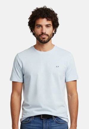 Uomo con capelli ricci e barba che indossa una t-shirt azzurro chiaro con piccolo logo sul petto, abbinata a jeans blu e cintura nera.