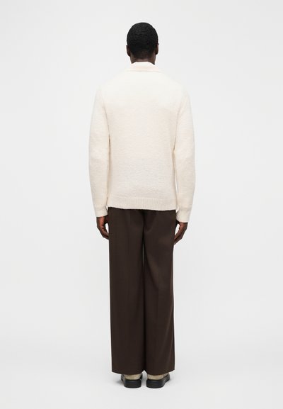 Cream-colored textured cardigan with a relaxed fit, ribbed cuffs, and a shawl collar, paired with brown wide-leg trousers.