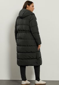 Parka negra larga y acolchada con capucha. Presenta detalles de cintura elástica y aberturas laterales para facilitar el movimiento. Acabado suave y mate.