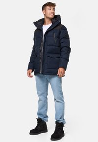 Mørkeblå puffjakke med hætte, quiltet design, frontlukning med lynlås og knaplommer, samt sorte detaljer på skuldrene. Sammen med lyseblå jeans og sorte støvler.