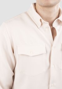 Lys beige skjorte med knapper, lavet af glat stof, med klassisk krave og to brystlommer med knaplukninger.