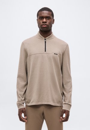 Giovane uomo che indossa un maglione beige con zip e il logo "BOSS" sul petto e pantaloni beige abbinati, in piedi davanti a uno sfondo chiaro e uniforme.