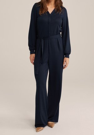 Nainen, jolla on päällään tummansininen pitkähihaisia haalarihousut, joissa on napit edessä, kangasvyö ja leveät lahkeet, yhdistettynä beigen värisiin teräväkärkisiin kenkiin, seisomassa yksiväristä taustaa vasten.