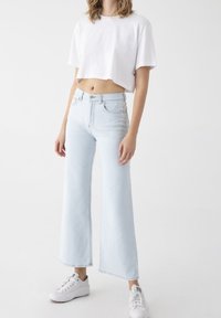 Jeans de pierna ancha de un azul claro con una cintura alta, combinados con una camiseta blanca corta. Zapatillas blancas completan el look.