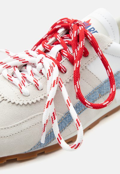 Világosszürke sportcipő piros-fehér mintás fűzőkkel, kék farmer oldalsávval és világosbarna gumitalppal, közelről bemutatva.