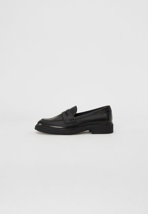 Mocassin en cuir noir avec un petit talon, un bout arrondi et un détail classique de bride à pampille, présenté en profil sur un fond uni.