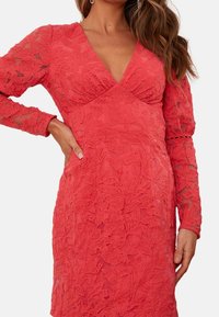 Röd spetsklänning med långa ärmar, djup V-ringning, blommönster, strukturerad yta, figursydd silhuett och intrikata detaljer på ärmarna.