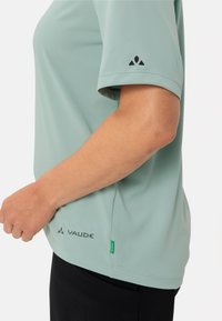 Chemise verte claire à manches courtes en tissu texturé et respirant. Présente le logo VAUDE en noir et une étiquette verte d'accent au bas.