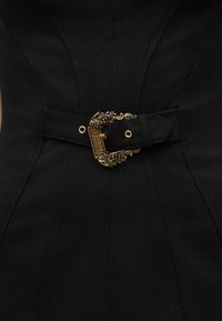 Μαύρο εφαρμοστό ένδυμα με φαρδιά υφασμάτινη ζώνη που διαθέτει περίτεχνη χρυσή αγκράφα χαραγμένη με την επιγραφή "Versace Jeans Couture".