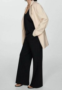 Blazer beige à fines rayures porté sur un débardeur noir, associé à un pantalon noir à jambes larges et des sandales noires. Textures lisses et coupe décontractée.