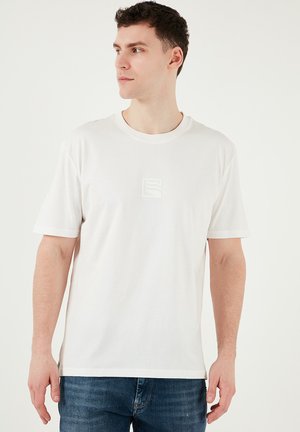Hvid bomulds T-shirt med korte ærmer, rund halsudskæring og et lille vævet logo på brystet. Kombineret med blå denim jeans.