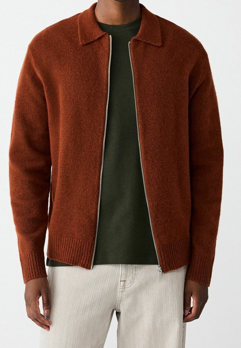 Homme portant un cardigan en laine couleur rouille avec fermeture éclair par-dessus une chemise vert foncé et un pantalon beige clair, debout devant un fond uni clair.