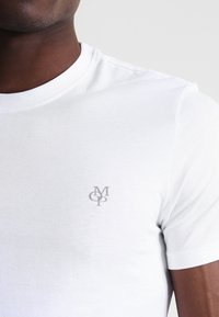 Camiseta blanca de algodón con cuello redondo y mangas cortas, con un pequeño logo gris en el lado izquierdo del pecho. Textura suave.