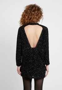 Svart paljettklänning med långa ärmar och en djup V-formad ryggdesign. Har en texturerad yta och en lös passform, som når midjan på låren.
