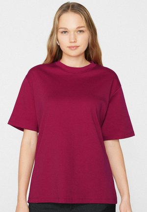 Jeune femme aux longs cheveux blonds portant un t-shirt uni à manches courtes magenta sur un fond blanc.