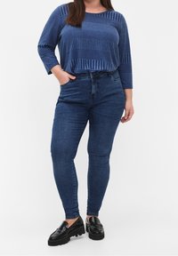 Frau in einem blauen gestreiften Langarmoberteil, dunkelbblauen Skinny-Jeans und schwarzen klobigen Slippern, die mit einer Hand in der Hosentasche steht.