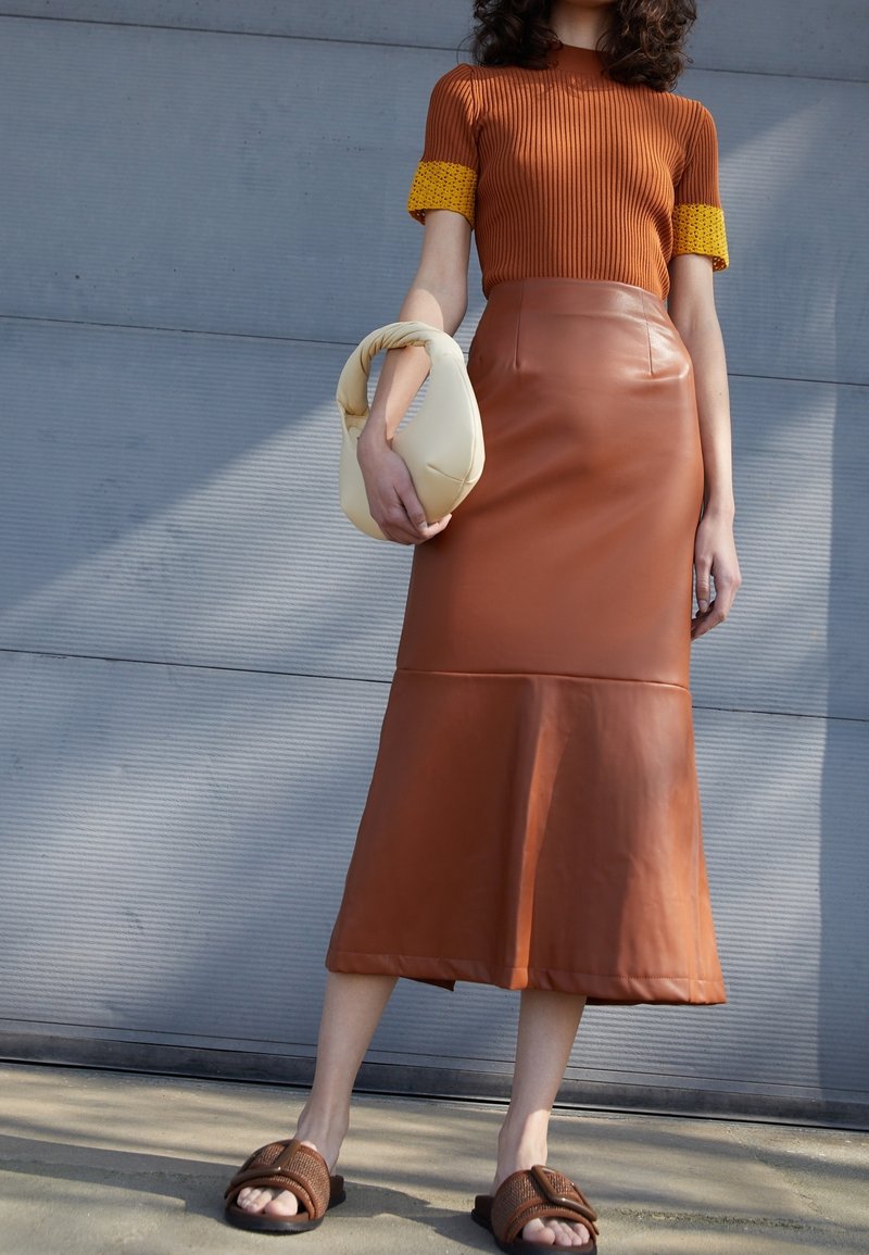 Haut en tricot à côtes marron avec des poignets à motifs jaunes, associé à une jupe midi en cuir marron et des sandales à lanière beiges. Tenant un sac crème.