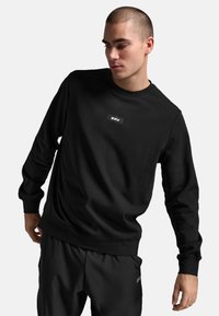 Sort sweatshirt lavet af et blødt materiale, med rund halsudskæring, lange ærmer og et lille logo-patch på brystet.