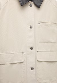 Beige jas met een zwarte leren kraag, voorzien van zilveren drukknopen en twee voorzakken met kleppen. Gladde stoftextuur.