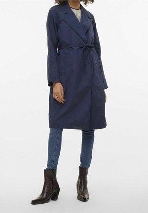 Kvinde iført en navyblå trenchcoat med bælte, stribet skjorte, blå jeans og mørkebrune ankelstøvler med hæl mod en ensartet hvid baggrund.