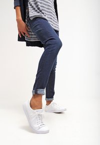 Zapatillas de malla blancas con cordones, combinadas con unos vaqueros oscuros ajustados con puños. El conjunto presenta una blusa de capas con rayas horizontales en blanco y azul marino.