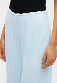Pantalon bleu clair à côtes avec bordure en dentelle blanche et petit nœud à la taille, porté avec un haut noir, main reposant légèrement sur la cuisse.