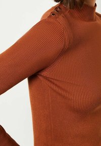 Maglione marrone a coste con collo alto e dettagli con bottoni sulle spalle. Il tessuto presenta una leggera texture; la forma aderente esalta la silhouette.