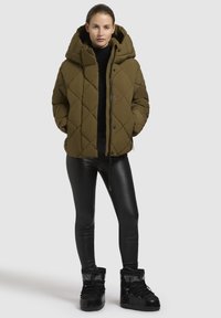 Vrouw in een olijfgroene gewatteerde pufferjack, zwarte turtleneck, glanzende zwarte leggings en zwarte winterlaarzen, staand met haar handen in de zakken.